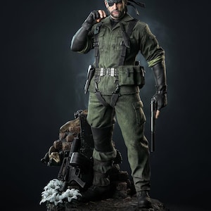 Könnte beinhalten: Eine detaillierte Sammlerfigur eines Soldaten in grüner taktischer Uniform, schwarzen Handschuhen und Augenklappe. Die Figur hält eine Pistole mit Schalldämpfer und steht auf einem Sockel mit Gewehr, Ziegeltrümmern und weißen Blumen. Text Tanuki Figures.