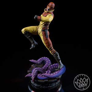 May include: A dynamic figurine of a character in a yellow and brown suit with a matching helmet and goggles, captured in a mid-air kicking pose. The figure stands atop a circular base featuring textured, purple octopus tentacles with visible suckers.