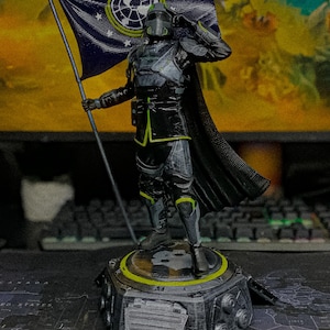 Puede incluir: Una figura detallada de un soldado con armadura negra y gris con detalles en verde neón, sobre una base hexagonal. El soldado sostiene una bandera azul oscuro con un emblema circular blanco y estrellas, mientras saluda con una mano.