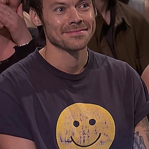 May include: A person wearing a dark grey t-shirt featuring a distressed yellow smiley face and the text "I hate you". The person has a tattoo on their arm that says "Slinky".