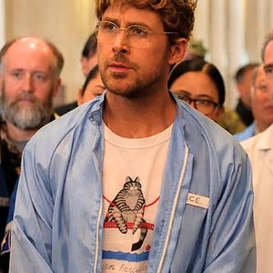 May include: A light blue zip-up jacket over a white t-shirt with a cat graphic. The t-shirt features a striped cat sitting on a red rope with the text "Sm Francisco" below. The person is wearing gold-rimmed glasses.