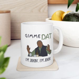 May include: White ceramic mug with the text "GIMME DAT" and "I'M JOKIN', I'M JOKIN'". The mug features a cartoon illustration of a man with his hands up, a bottle, and a wine glass. A fun, novelty item.