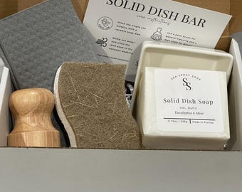 May include: A dish cleaning kit in a cardboard box containing a solid dish soap bar labeled Sea Spray Soap Eucalyptus & Mint 11.75oz 333g, a white ceramic soap dish, a wooden palm brush, a brown fibrous scrubbing sponge, and a gray cleaning cloth.