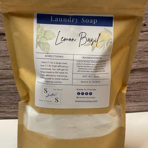 Op de afbeelding: Een bruine papieren zak met Lemon Basil wasmiddel. Het etiket bevat de tekst "Laundry Soap" en "Lemon Basil". De ingredi&euml;nten en instructies staan vermeld. Het nettogewicht is 907g. De merknaam is Sea Spray Soap Company.