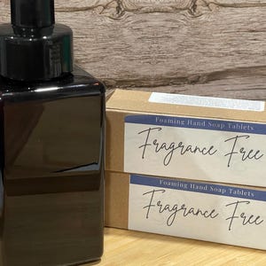 May include: A dark brown square plastic soap dispenser with a black pump mechanism positioned next to two rectangular cardboard boxes labeled Foaming Hand Soap Tablets Fragrance Free in black script text on a white background label.