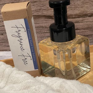 May include: A square clear glass foaming hand soap dispenser with a black pump mechanism, accompanied by a rectangular cardboard box labeled Fragrance Free and Foaming Hand Soap Tablets, positioned next to a white textured fabric.