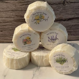 May include: A stack of six round, wrapped solid dish soap refills. Each soap is labeled with a different scent: Lemon Basil, Citrus, Lavender, and Lemongrass. The labels also include the words "Sea Spray Soap" and "Solid Dish Soap Refill, Net 6 oz (170g)."