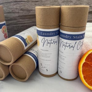 Op de afbeelding: Verschillende cilindrische kartonnen containers met vlekverwijderaar voor de was worden getoond, met labels met de tekst "Laundry Stain Natural Citrus". De containers zijn bruin met witte en blauwe labels en een gesneden sinaasappel is zichtbaar.