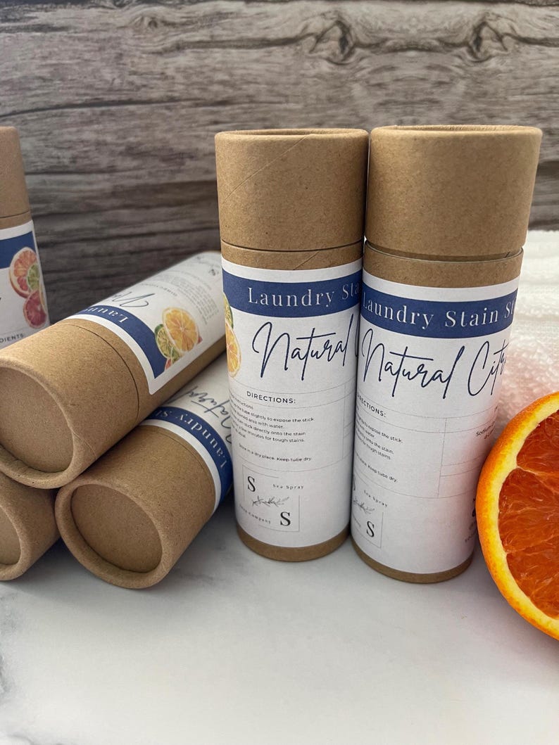 Op de afbeelding: Verschillende cilindrische kartonnen containers met vlekverwijderaar voor de was worden getoond, met labels met de tekst "Laundry Stain Natural Citrus". De containers zijn bruin met witte en blauwe labels en een gesneden sinaasappel is zichtbaar.