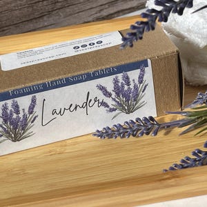 Op de afbeelding: Een bruine kartonnen doos met het opschrift "Foaming Hand Soap Tablets" en een lavendelontwerp. Het etiket leest "Lavender" in script. De doos staat op een houten dienblad met lavendeltakken en een witte handdoek op de achtergrond.