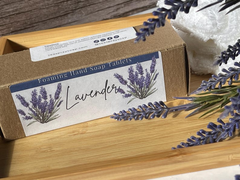 Op de afbeelding: Een bruine kartonnen doos met het opschrift "Foaming Hand Soap Tablets" en een lavendelontwerp. Het etiket leest "Lavender" in script. De doos staat op een houten dienblad met lavendeltakken en een witte handdoek op de achtergrond.
