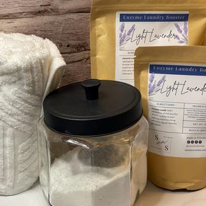 Op de afbeelding: Een heldere glazen pot met een zwarte deksel gevuld met wit poeder, naast twee bruine papieren zakken met het opschrift "Enzyme Laundry Booster Light Lavender". Een witte handdoek is gedeeltelijk zichtbaar aan de linkerkant.