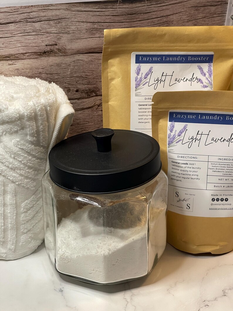 Op de afbeelding: Een heldere glazen pot met een zwarte deksel gevuld met wit poeder, naast twee bruine papieren zakken met het opschrift "Enzyme Laundry Booster Light Lavender". Een witte handdoek is gedeeltelijk zichtbaar aan de linkerkant.
