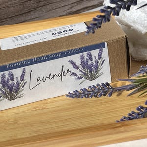 May include: A rectangular brown cardboard box of foaming hand soap tablets with a white label featuring purple lavender illustrations and the text Foaming Hand Soap Tablets Lavender. The box includes social media icons and a website address.