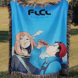 Peut inclure: Un plaid tissé avec un fond bleu vif et le texte FLCL et FOOLY COOLY en haut. Le motif représente un personnage de style anime aux cheveux roses à côté d'un personnage en veste bleue et casquette rouge buvant dans une canette Indigo.