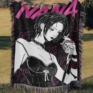 May include: Black woven throw blanket with a pink and white graphic of a woman holding a microphone and cigarette. The word "NANA" is in pink at the top. The blanket has a fringed edge.