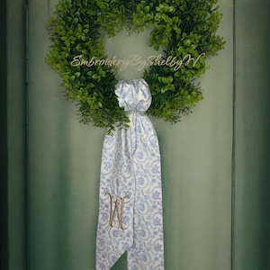 May include: A green wreath with a white and blue ribbon with a monogram "W" embroidered on it. The ribbon is tied in a bow. The wreath is hanging on a green door. The text "Embroidery By Shelby W" is visible on the wreath.