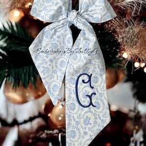 May include: A white and blue patterned bow with a blue embroidered letter "C" hanging on a Christmas tree. The bow is tied with a knot and has a long tail. The text "Embroidery By Shelby G" is visible on the bow.