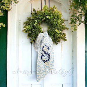 May include: A white door with a green wreath and a blue and white ribbon with an embroidered letter "S" hanging on it. The text "Antiques by G" is visible on the door.