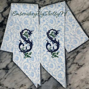 May include: Two white and light blue floral patterned sashes with a dark blue embroidered letter "S" and green accents. The text "Embroidery By Shelby W" is written in green cursive across the top.