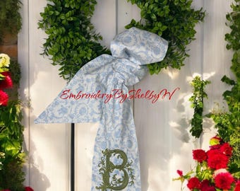 May include: A green wreath with a blue and white patterned fabric bow tied around it. The bow has a green embroidered letter "B" in the center. The wreath is hanging on a white wooden door.
