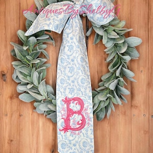 May include: A green leafy wreath with a blue and white patterned ribbon tied in a bow. The ribbon has a pink embroidered letter "B" in a decorative font.
