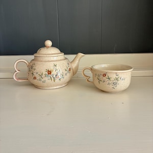 May include: A ceramic teapot with a lid and a matching teacup, both featuring a cream-colored speckled finish and a delicate floral pattern in shades of blue, orange, and green, with distinctive heart-shaped handles on both pieces.