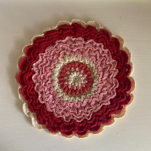 May include: A handmade, circular crochet doily featuring concentric rings of color. The center is a deep red, transitioning through shades of pink and cream, with a scalloped edge. The doily is likely used as a decorative item.