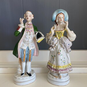 May include: A pair of vintage porcelain figurines depicting a man and a woman in 18th-century attire. The man is wearing a green jacket with gold trim, a blue and white striped vest, and white breeches. The woman is wearing a white dress with a yellow bodice, a blue hat, and a floral print.