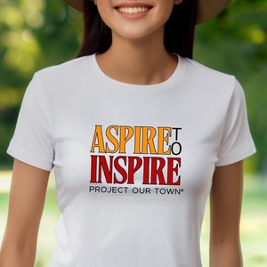 Peut inclure: T-shirt blanc avec un graphisme rouge et orange qui dit "ASPIRE TO INSPIRE" et "PROJECT OUR TOWN*"