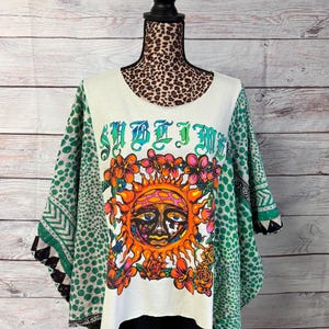 May include: A white short-sleeved top featuring a graphic of a sun with a face surrounded by orange and pink flowers. The text SUBLIME is printed in green above the sun. The sleeves have a green and white polka dot and geometric pattern.