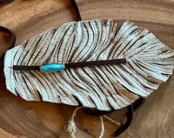 May include: White leather feather necklace with a turquoise stone accent. The necklace is on a wooden surface with a tag that reads "By the Wind".