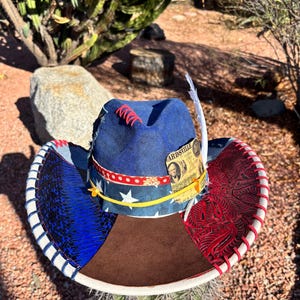May include: A unique cowboy hat with a blue crown, adorned with red stitching and a decorative feather. The brim features a patchwork design with denim, red patterned fabric, and a brown suede center. A yellow ribbon and a "Wardsville" label add detail.