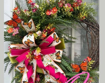 May include: A spring wreath with a pink and white ribbon with a butterfly pattern. The wreath is decorated with green leaves and orange butterflies. The wreath is made of twigs and has a natural look.