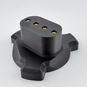 Puede incluir: Un componente de base negro mate de tres puntas con una sección superior elevada de forma rectangular redondeada. Esta sección superior contiene cuatro puntos de contacto metálicos de color dorado, circulares y espaciados uniformemente.