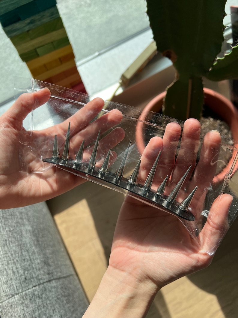 May include: A silver-colored hair comb with sharp, conical spikes. The comb is held in a clear plastic package. The spikes are evenly spaced along the comb's base. The background includes a window and a potted plant.