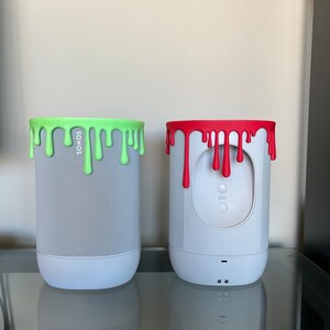 Könnte beinhalten: Zwei weiße Sonos-Lautsprecher mit einem grünen und roten tropfenden Farbmuster auf der Oberseite. Die Lautsprecher stehen auf einer Glasfläche.