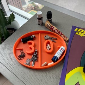 Può includere: Un vassoio ovale arancione con scomparti contiene vari oggetti. Sono visibili un accendino nero, chiavi e un tubetto di Aquaphor. Sono presenti anche piccole bottiglie, pillole, una catena, un ciondolo e un Raw Cone. I numeri "3" e "0" sono stampati sul vassoio.