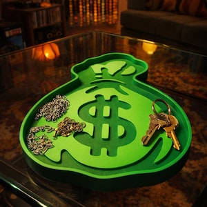 May include: A bright green tray shaped like a money bag with a large dollar sign cutout in the centre. The tray holds two thin metallic chains on the left side and a metal keyring with several keys on the right side of the dollar sign.