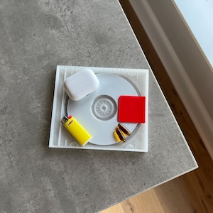 Puede incluir: Una colección de objetos cotidianos dispuestos sobre una superficie gris. Los elementos incluyen un CD blanco, una funda de auriculares inalámbricos blanca, un encendedor BIC amarillo, dos cápsulas marrones y un pequeño cuadrado rojo.