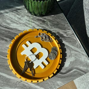 Puede incluir: Una bandeja circular amarilla con un símbolo de Bitcoin blanco en relieve en el centro. La bandeja tiene un borde texturizado y acanalado. Llaves y monedas descansan sobre la bandeja.