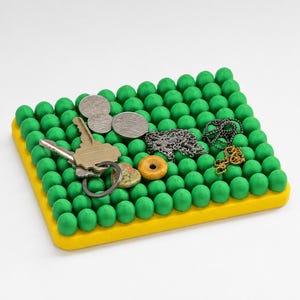 Op de afbeelding: Een gele en groene organizer met groene bollen, met sleutels, munten en sieraden. De organizer heeft een rechthoekige vorm met een gele rand en is gevuld met groene bollen.