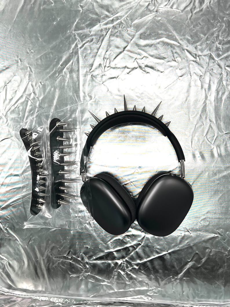 May include: Black over-ear headphones with silver spikes on the headband. A matching black hair comb with silver spikes is also visible. The items are displayed on a crinkled silver surface.