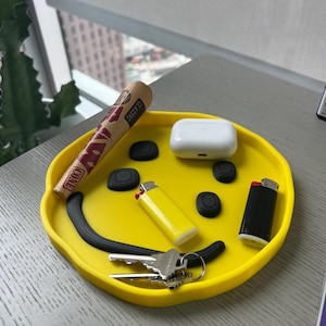 May include: A yellow smiley face silicone tray with a black outline. The tray contains a rolled up brown paper, a lighter, a set of keys, a black lighter, and a white wireless charging case.