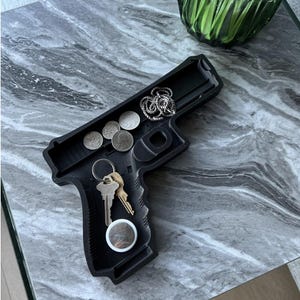 Puede incluir: Bandeja negra en forma de pistola con una cadena plateada, monedas y llaves en su interior. La bandeja está sobre una superficie de mármol gris.
