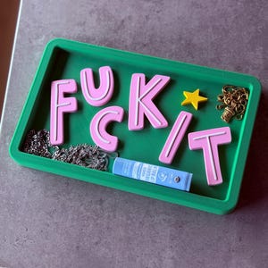 May include: A green rectangular tray with the word "F*CK IT" spelled out in pink letters with white outlines. The tray also contains a gold chain, a silver chain, a yellow star, and a blue lip balm tube.