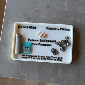 Könnte beinhalten: Eine weiße rechteckige Ablage mit einem Feuerzeug, Listerine, Pillen und Schmuck. Die Ablage trägt die Aufschrift "212 555 6342", "PIERCE & PIERCE", "PATRICK BATEMAN VICE PRESIDENT" und eine Adresse.