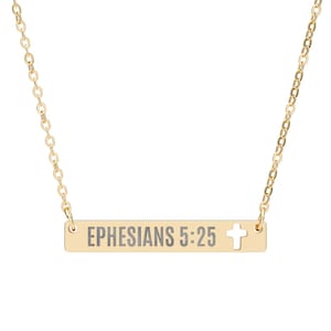 Puede incluir: Collar dorado con un colgante rectangular con la inscripción "EPHESIANS 5:25" y un recorte de cruz. La fina cadena se fija al colgante. Una pieza de joyería.