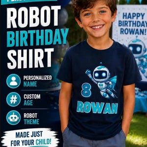 May include: A navy blue short-sleeved t-shirt featuring a graphic of a white and blue robot flying with a cape. The number 8 is printed in blue above the name ROWAN in large blue capital letters on the front of the shirt.