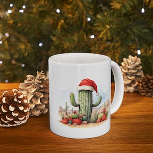 May include: White ceramic mug featuring a watercolor illustration of a cactus wearing a Santa hat. The design includes desert plants and a landscape with a blue sky and red flowers. The mug has a curved handle.
